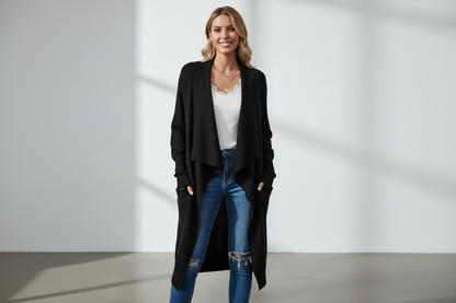Woman wearing a black cardigan, white shirt, and blue jeans standing against a plain background