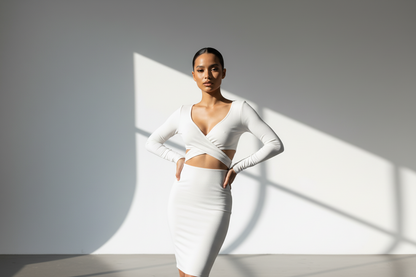 Woman wearing a white outfit standing against a gray wall with light casting shadows.