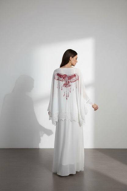 Woman wearing a white dress with red embroidery standing against a white wall.