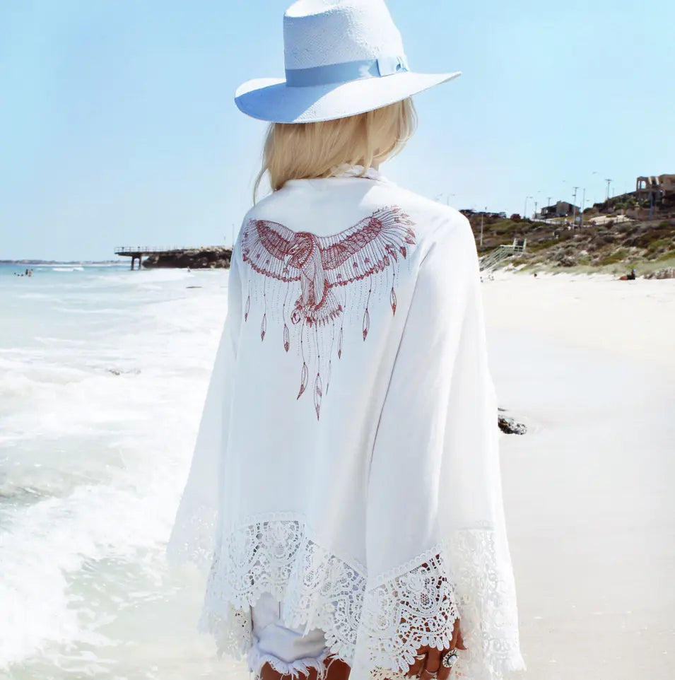 Person wearing a white lace cover-up with red embroidery on a beach.